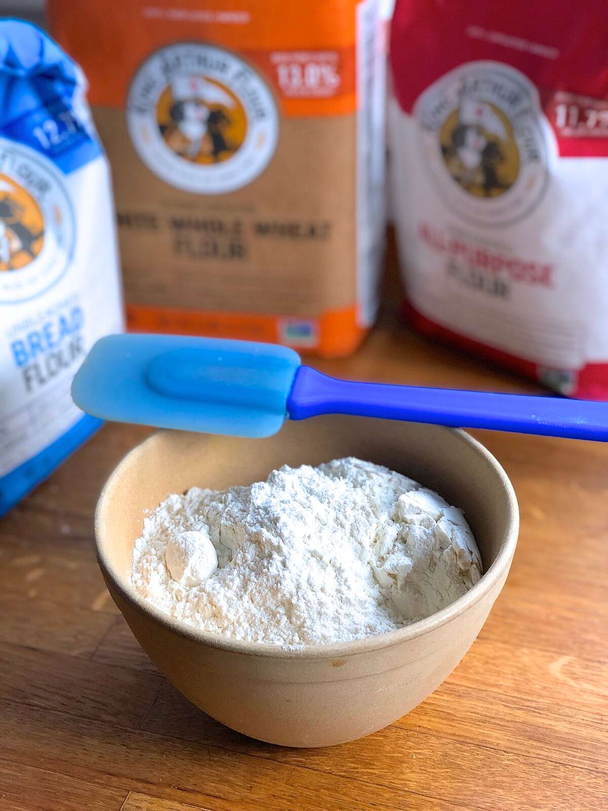 Bags of whole wheat, all-purpose, and bread flour with a bowl of all-purpose flour and mixing spoon.