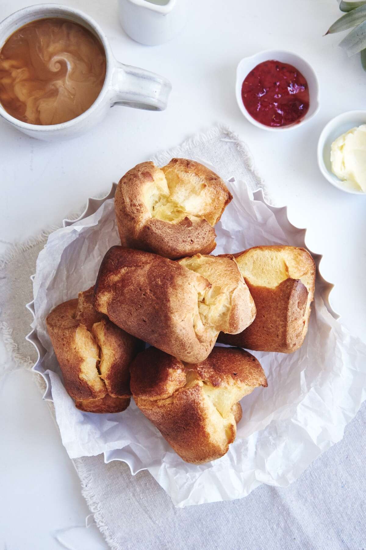 Basket of sourdough popovers on a table with jam, butter, and a latte.