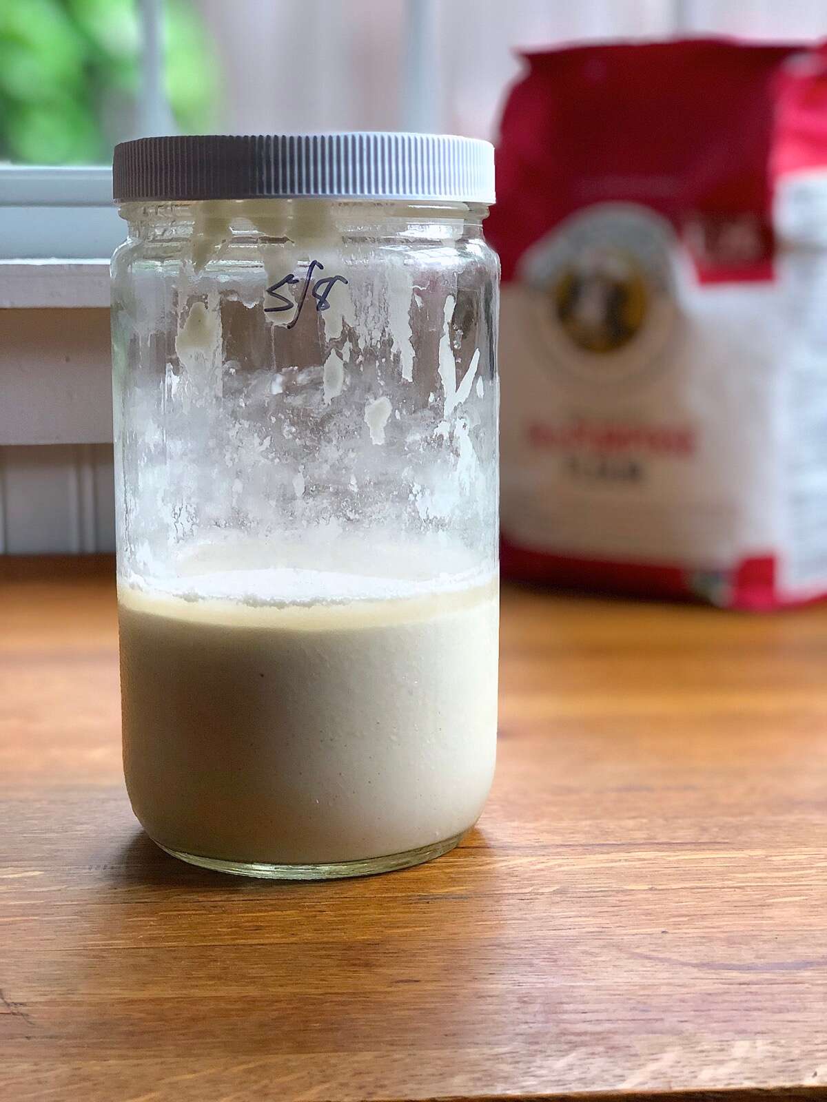 Sourdough starter in a straight-sided glass jar on a wooden table, bag of flour in the background.